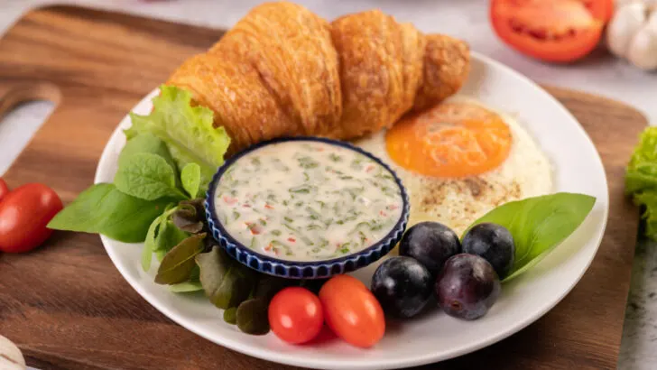 Breakfast consists of croissant, fried egg, salad dressing, black grapes, and tomatoes